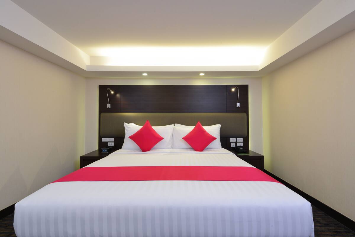 Une chambre d’hôtel avec un grand lit, deux tables de chevet, une tête de lit moderne et un coussin décoratif rouge.