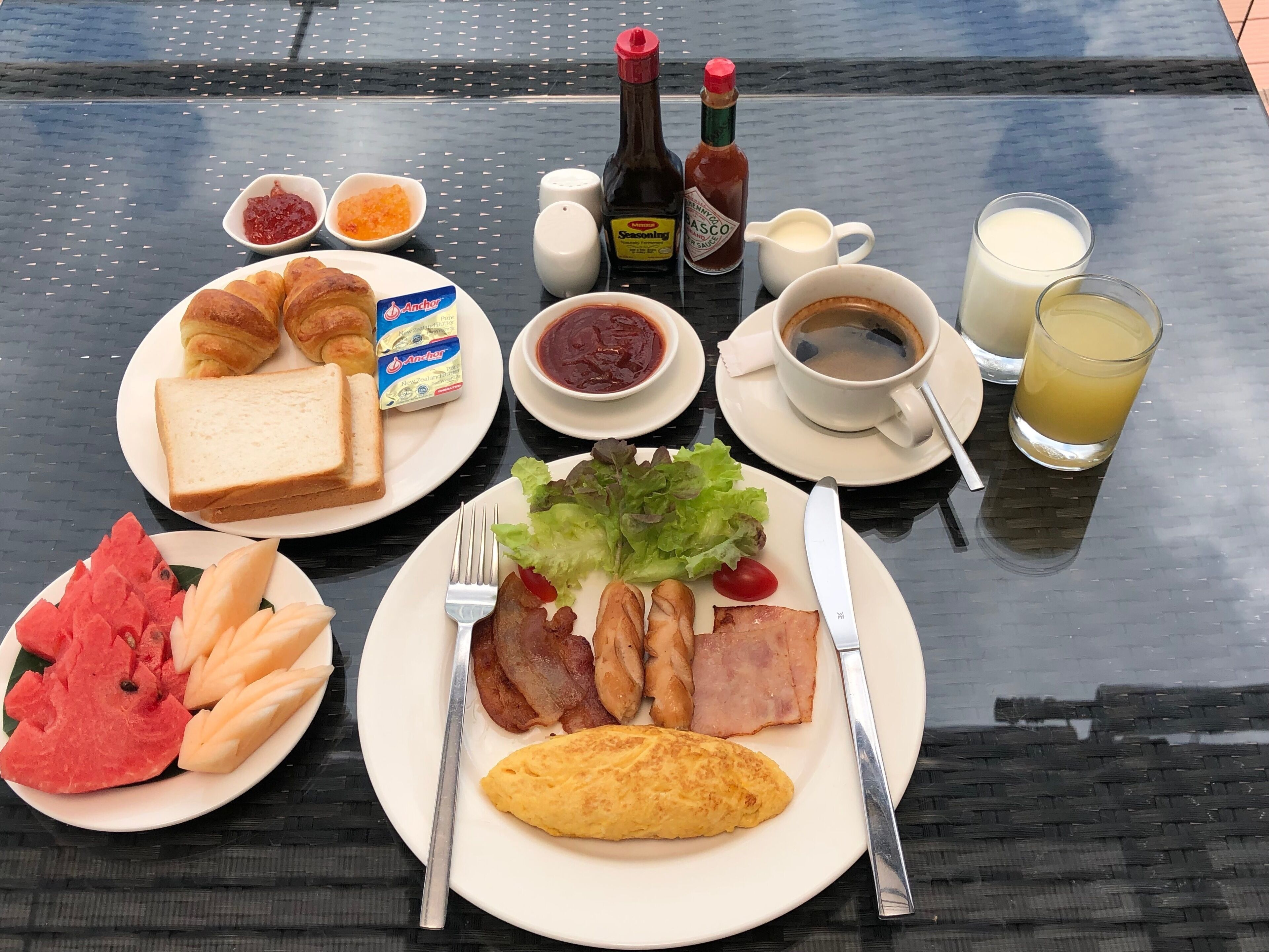 A breakfast table with croissants, toast, ham, eggs, and fruit. There are condiments, coffee, and a glass of juice.