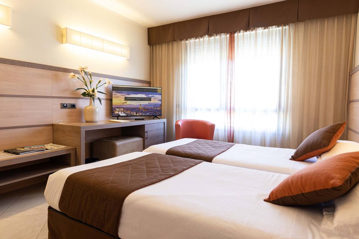 A hotel room with two beds, a desk with a computer, a vase with flowers, and a large window with curtains.