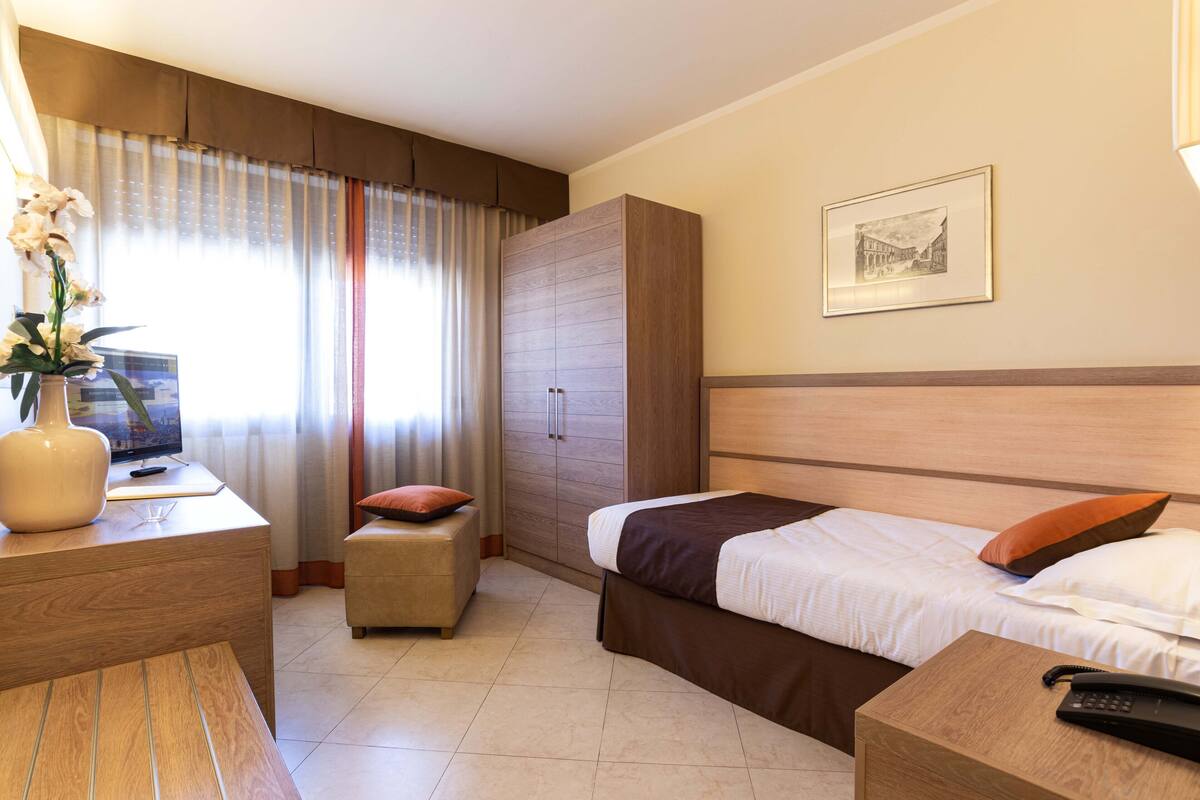 A hotel room with a bed, bedside table, a desk with a lamp, a chair, a wardrobe, and a window with curtains.