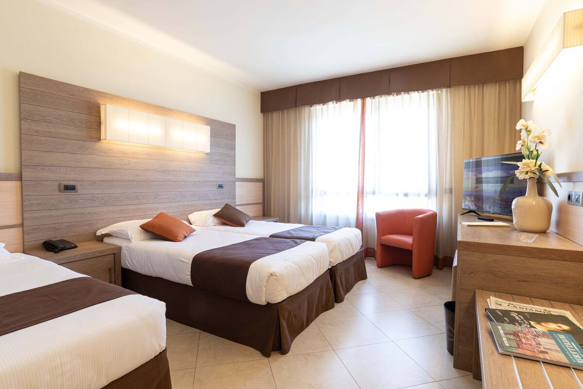 A hotel room with two beds, a desk with a TV, a chair, and a window with curtains.