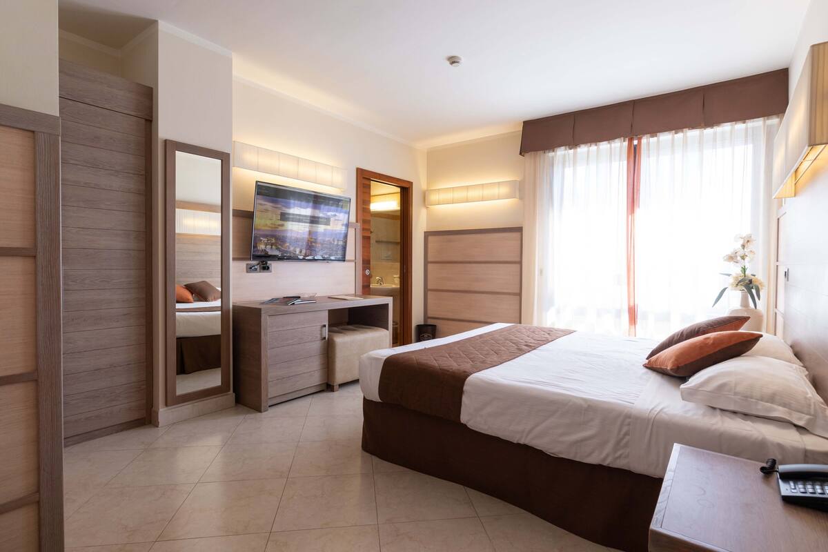 A modern hotel room with a large bed, a desk, a mirror, and a television.