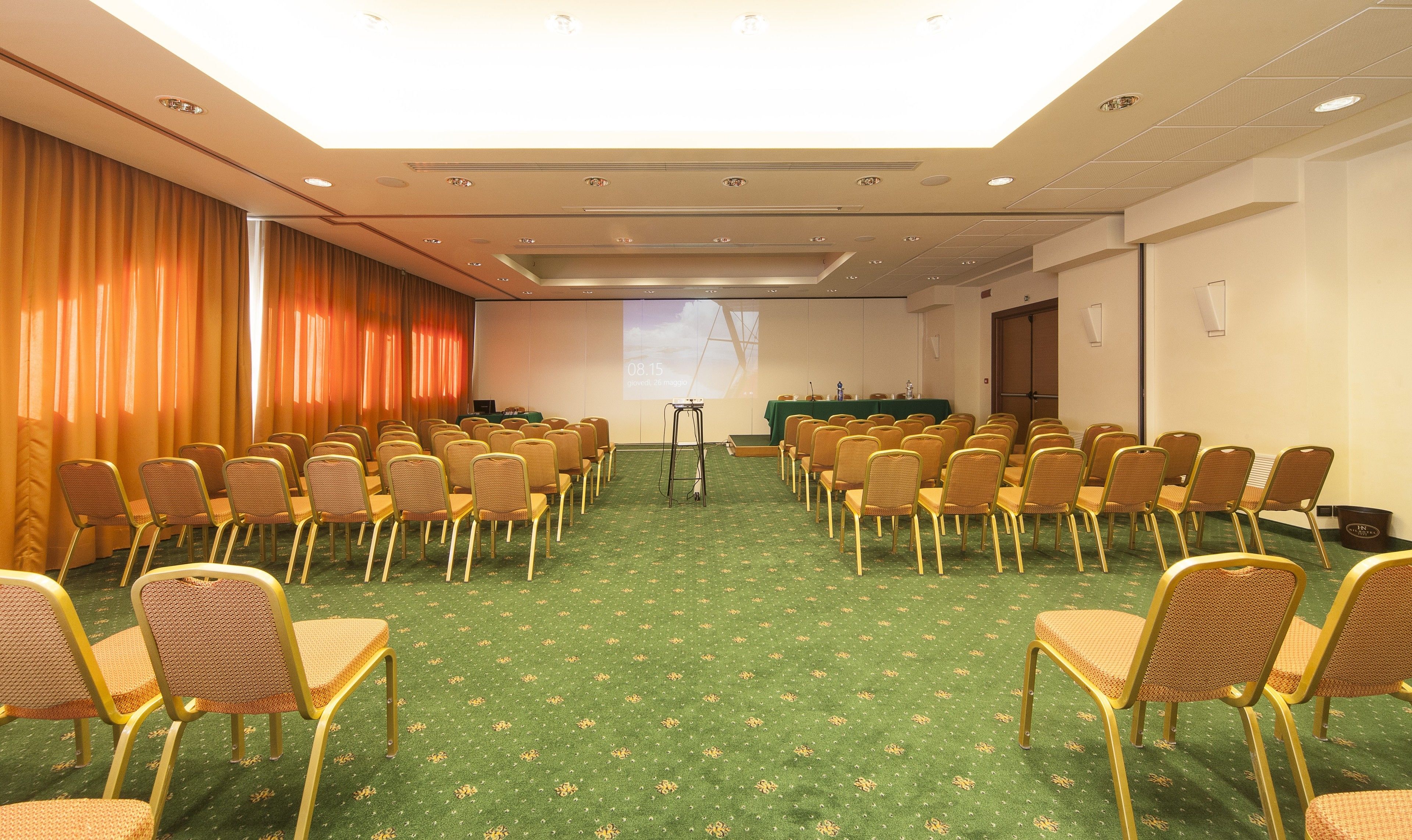 A conference room with rows of chairs facing a projection screen, a stage with a table, and a door on the right.