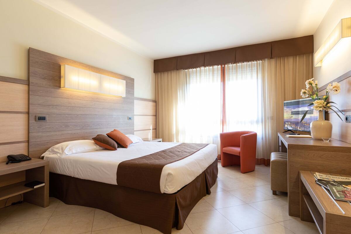 A hotel room with a large bed, a desk with a chair, a TV, and a window with curtains.