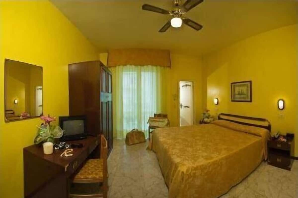 Ein Hotelzimmer mit einem Bett, einem Schreibtisch mit Computer, einem Sessel, einem Deckenventilator und einem Fenster mit Vorhängen.