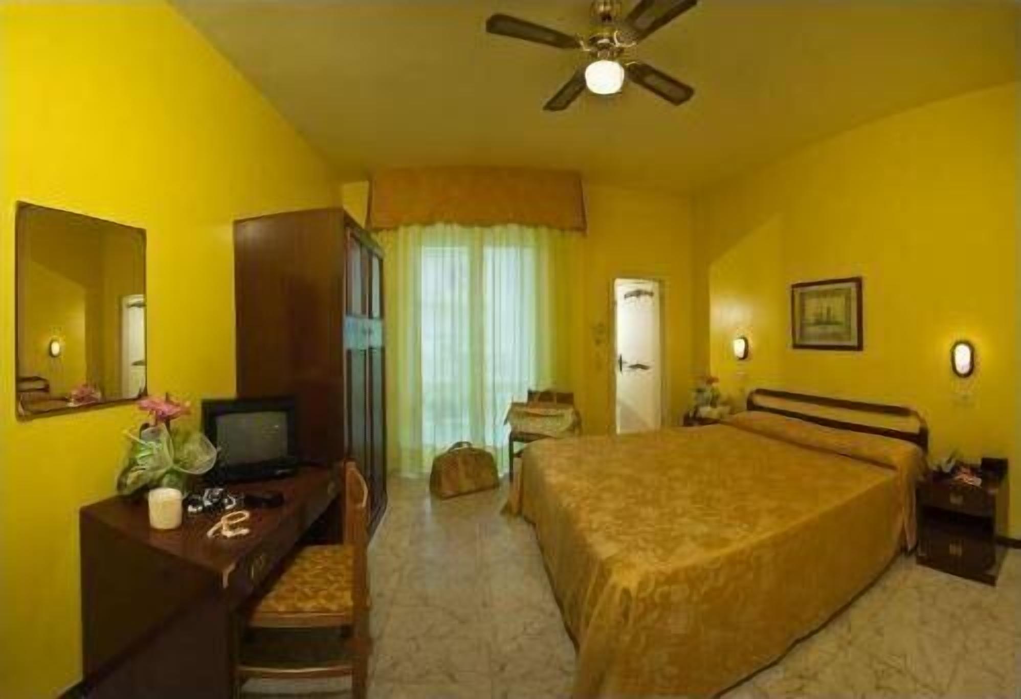 Habitación de hotel con una cama, un escritorio con ordenador, una silla, un ventilador de techo y una ventana con cortinas.