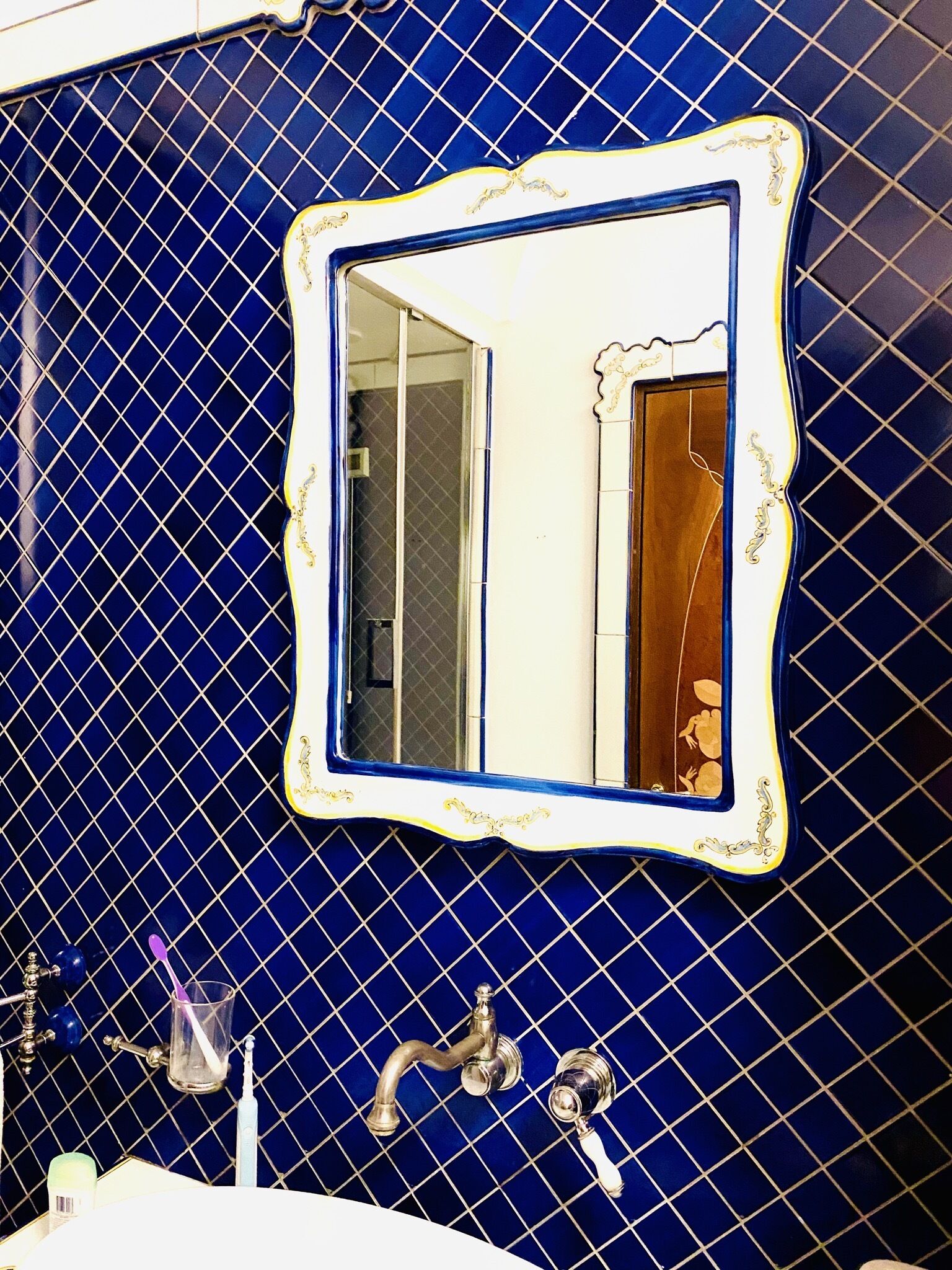 Un baño con pared de azulejos azules, espejo con marco blanco y dos grifos.