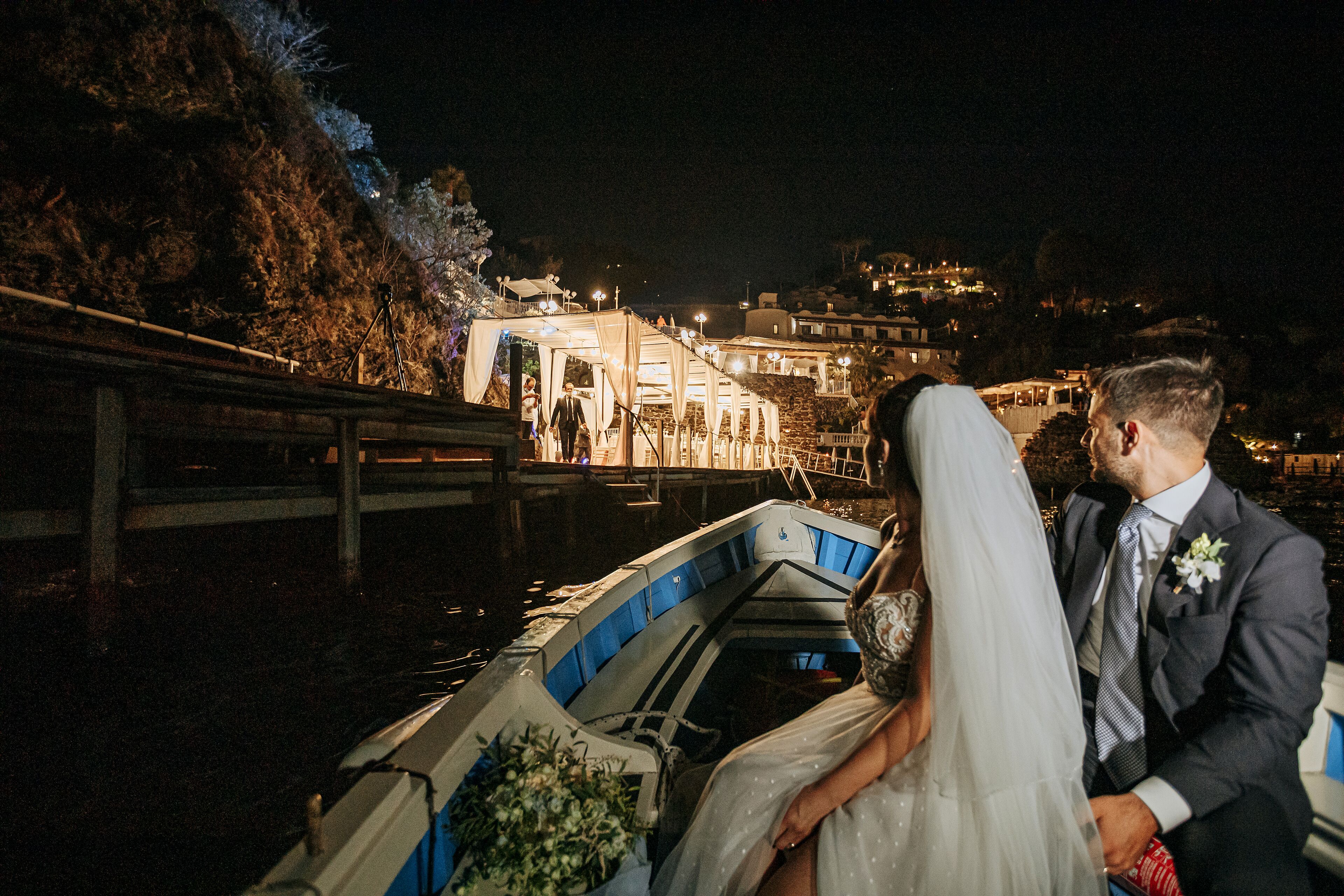 Una pareja de novios en un barco, con edificios iluminados y un puente como telón de fondo.