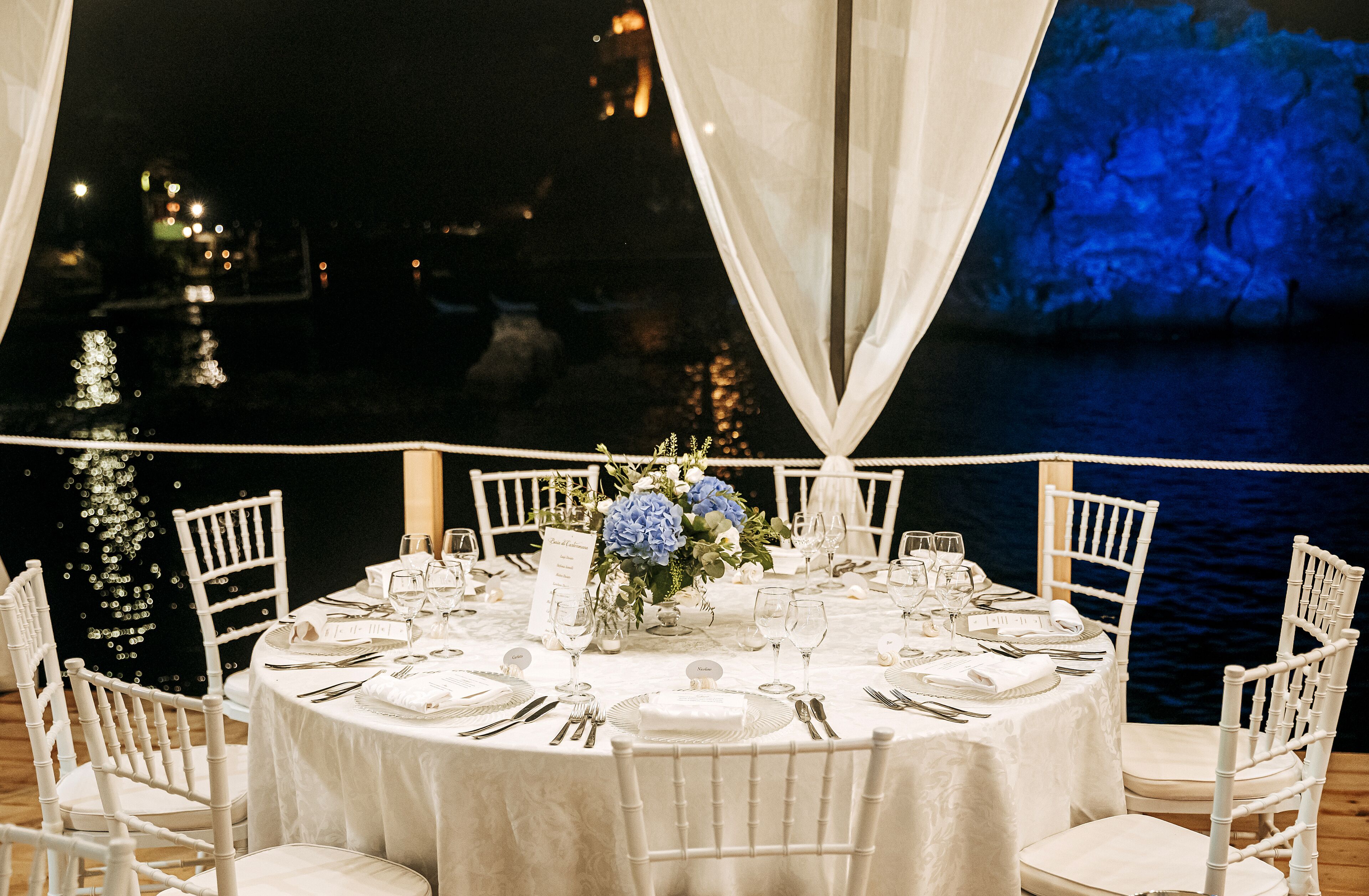 Una mesa redonda de comedor lista para un evento formal, con manteles y sillas blancas, y un centro de mesa floral.