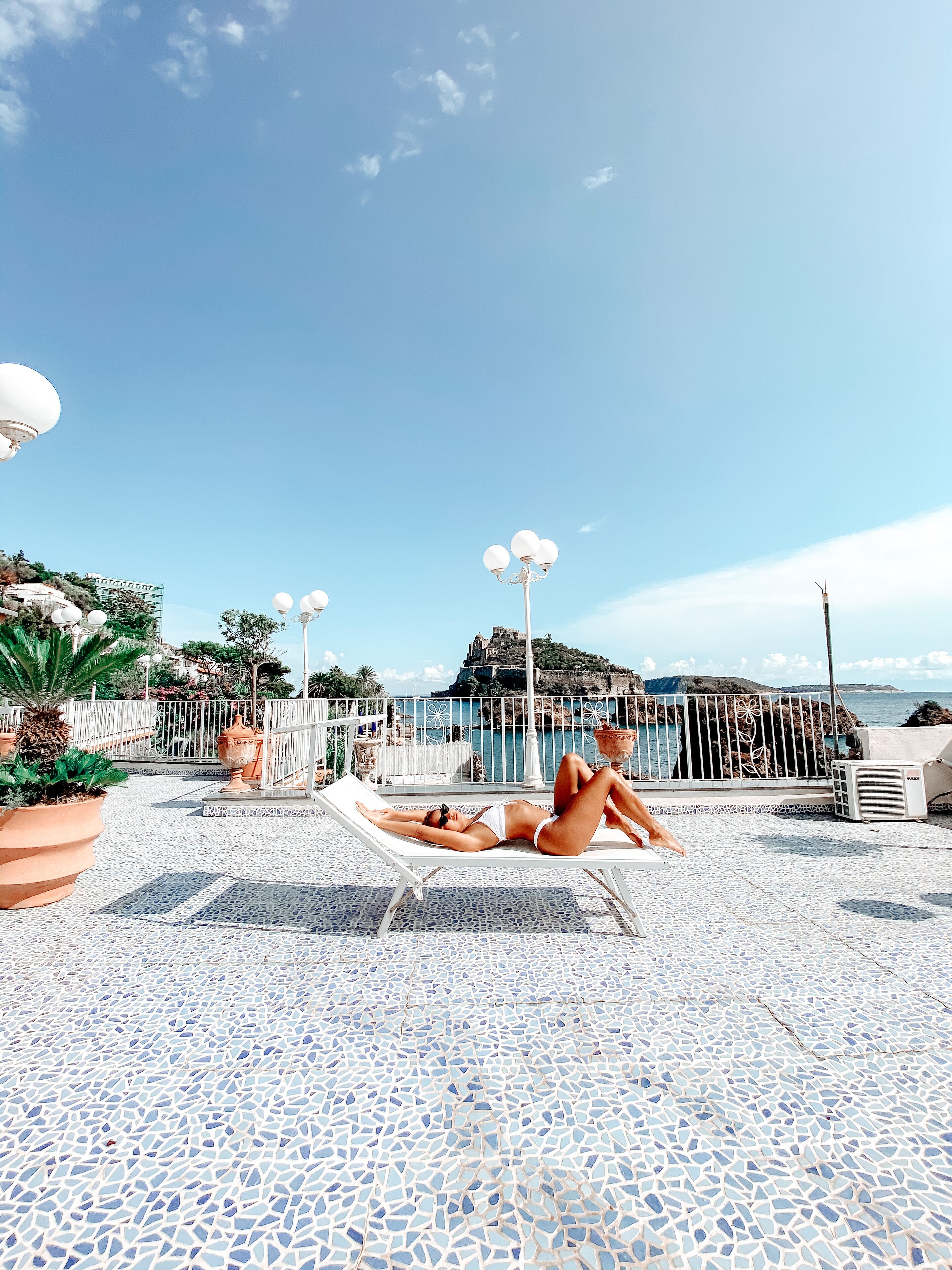 Un área al aire libre con baldosas, una piscina, sillas de descanso y vistas al mar y una isla distante.