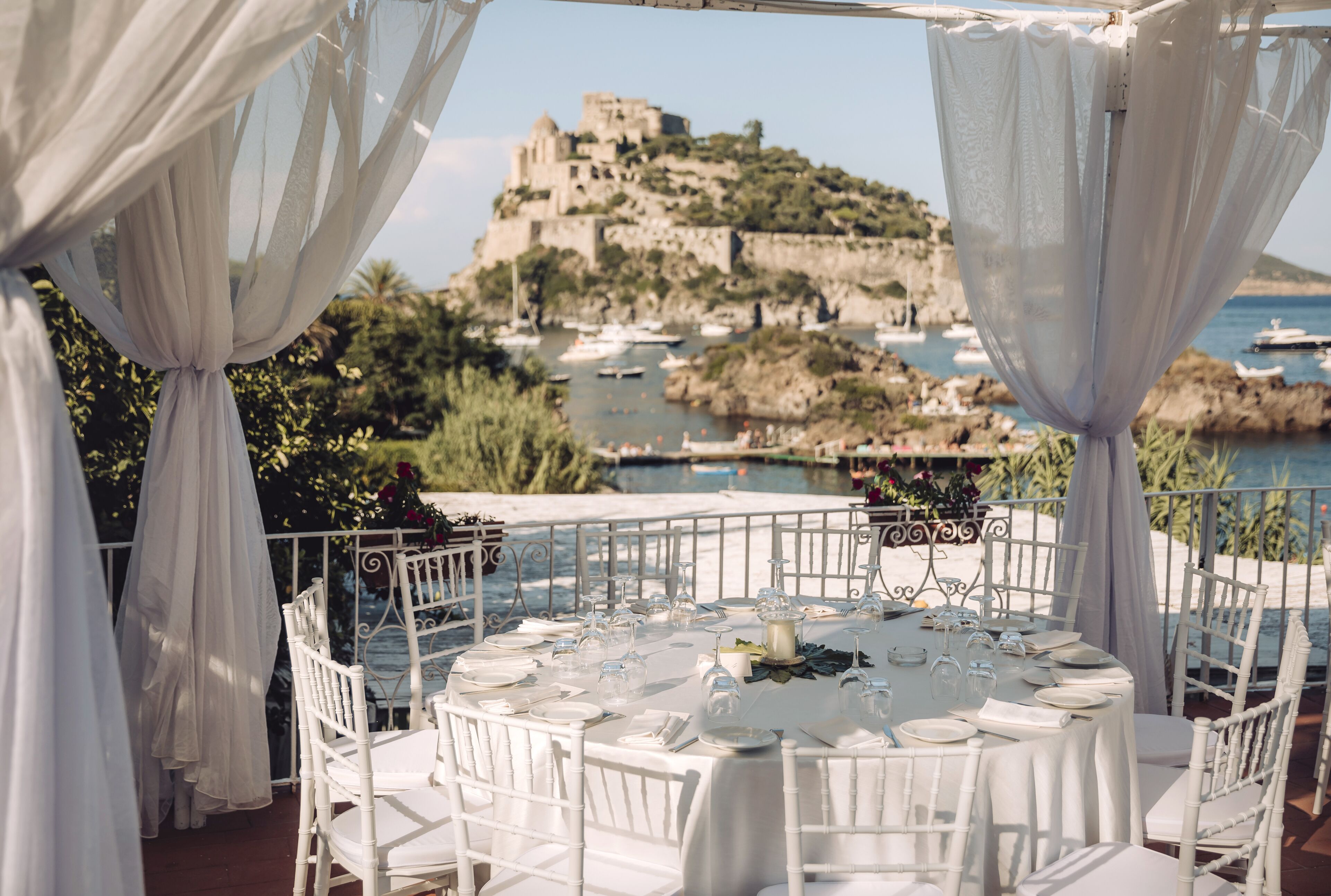 An outdoor dining area with white tables and chairs, set against a scenic backdrop of a coastal landscape with boats and a castle.