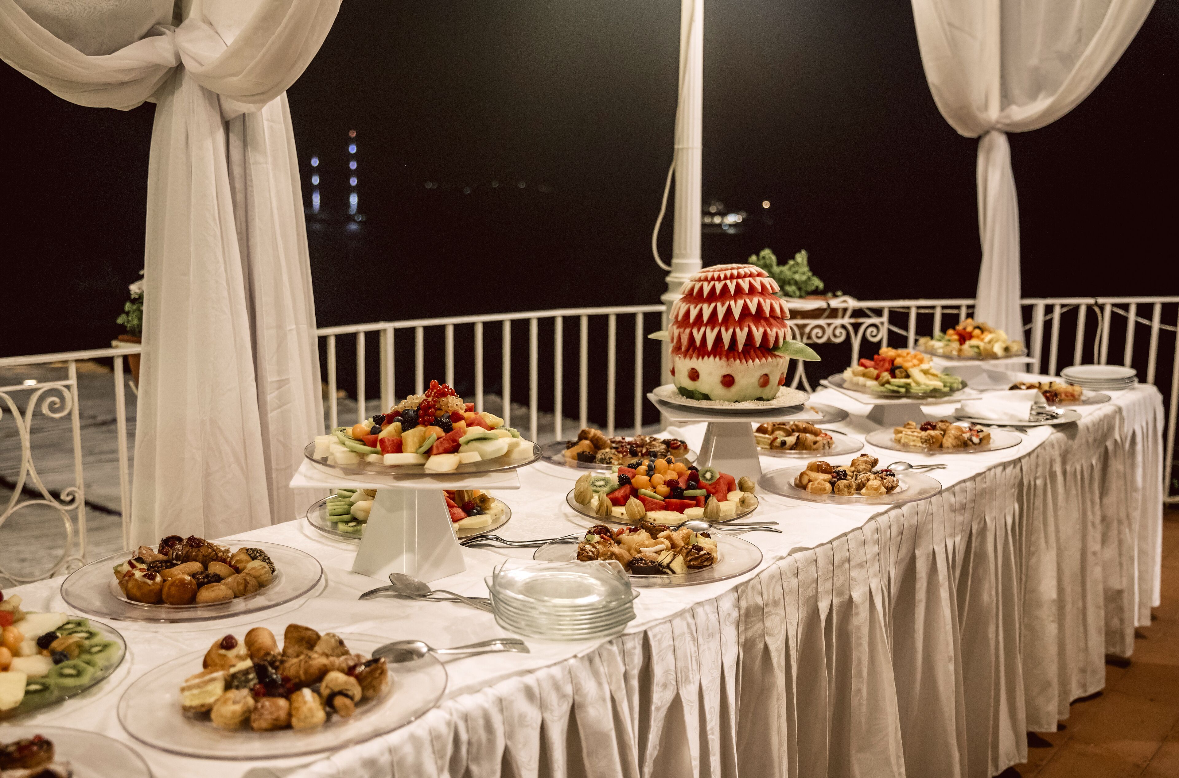 Une table de buffet garnie de divers plats et d’un gâteau, installée à l’extérieur, avec des draperies blanches et une balustrade en arrière-plan.
