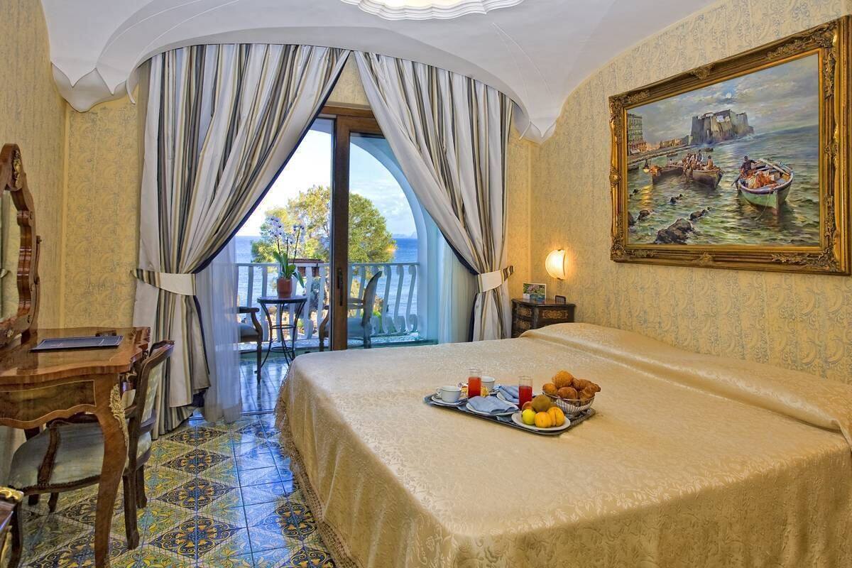 A hotel room with a bed, breakfast tray, curtains, a painting, a table, and a view of the sea.