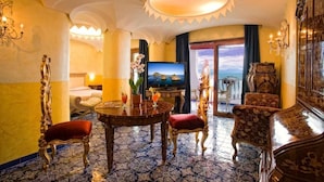 Quarto de hotel luxuoso com uma cama grande, televisão, mesa de jantar com cadeiras e vista para o mar através de uma janela ampla.