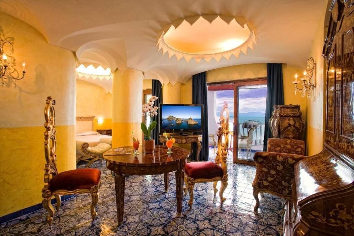 A luxurious hotel room with a large bed, a television, a dining table with chairs, and a view of the sea through a large window.