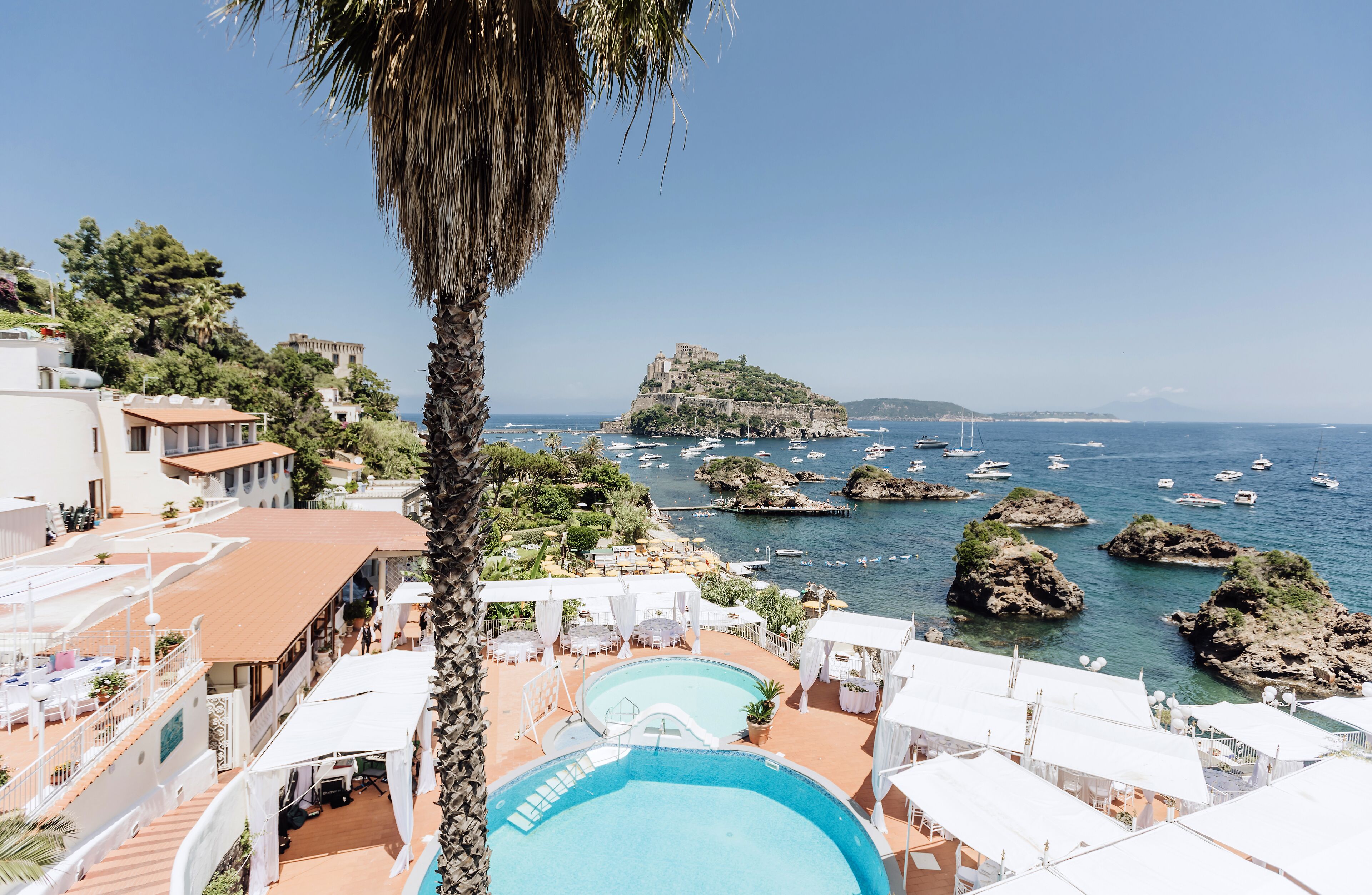 A resort with a pool area, white canopied seating, and a view of the sea and rocky outcrops.