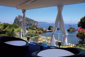 Una terraza con mesas al aire libre, vista al mar y un castillo al fondo.