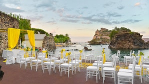 Decoração de casamento ao ar livre com cadeiras brancas e detalhes amarelos, com vista para um corpo de água e um edifício histórico.