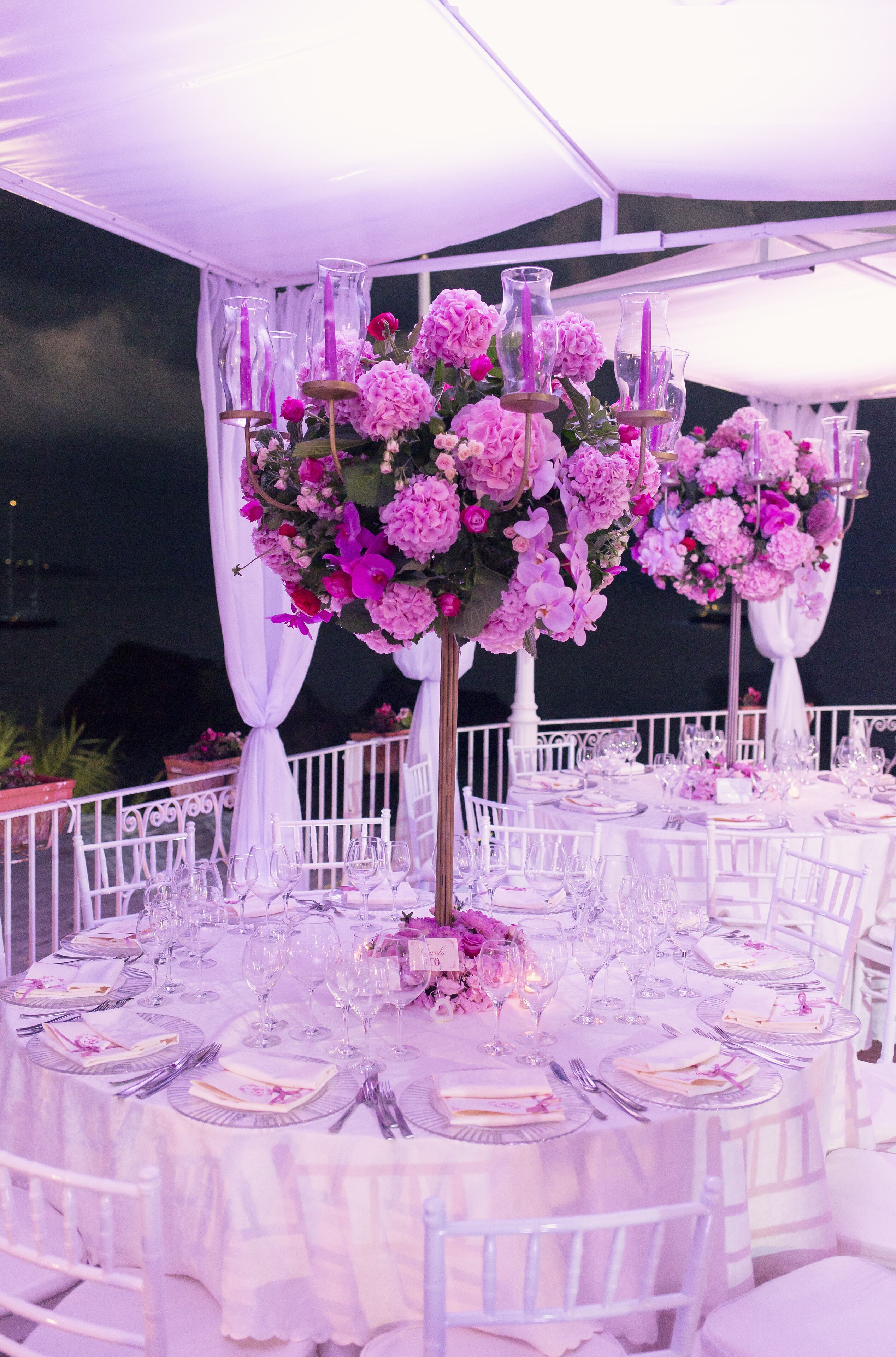 A round table set with white chairs, surrounded by white drapes and pink flowers.