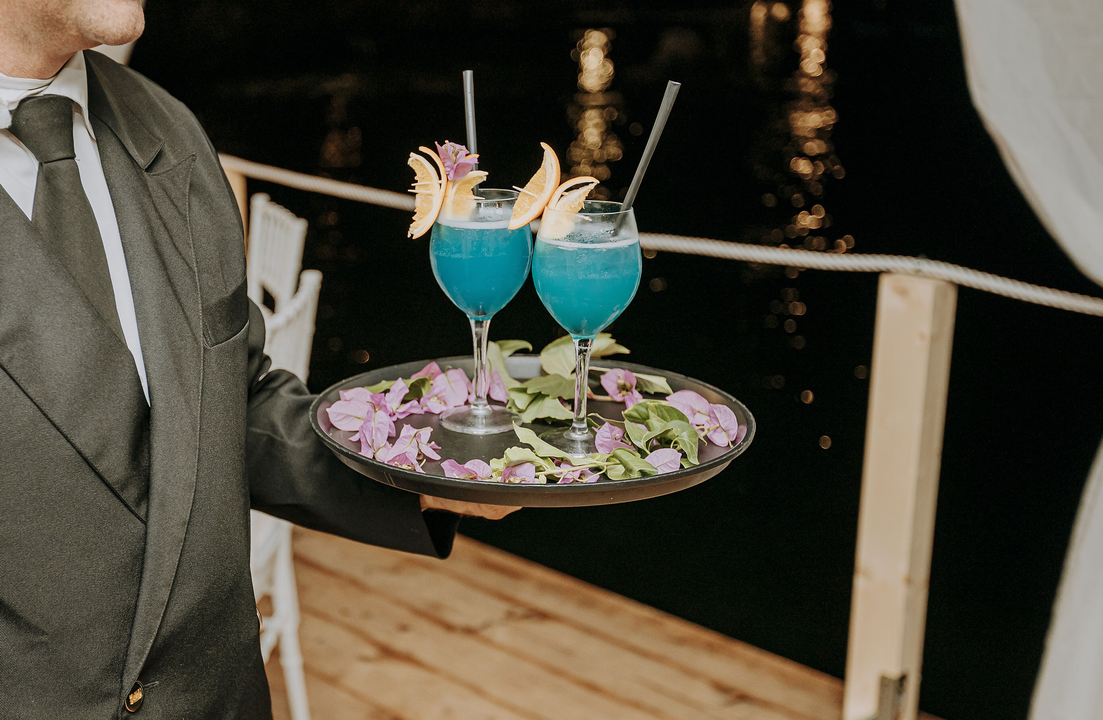 A person holding a tray with two blue cocktails garnished with fruit and flowers, set against a nighttime backdrop with a railing and lights.