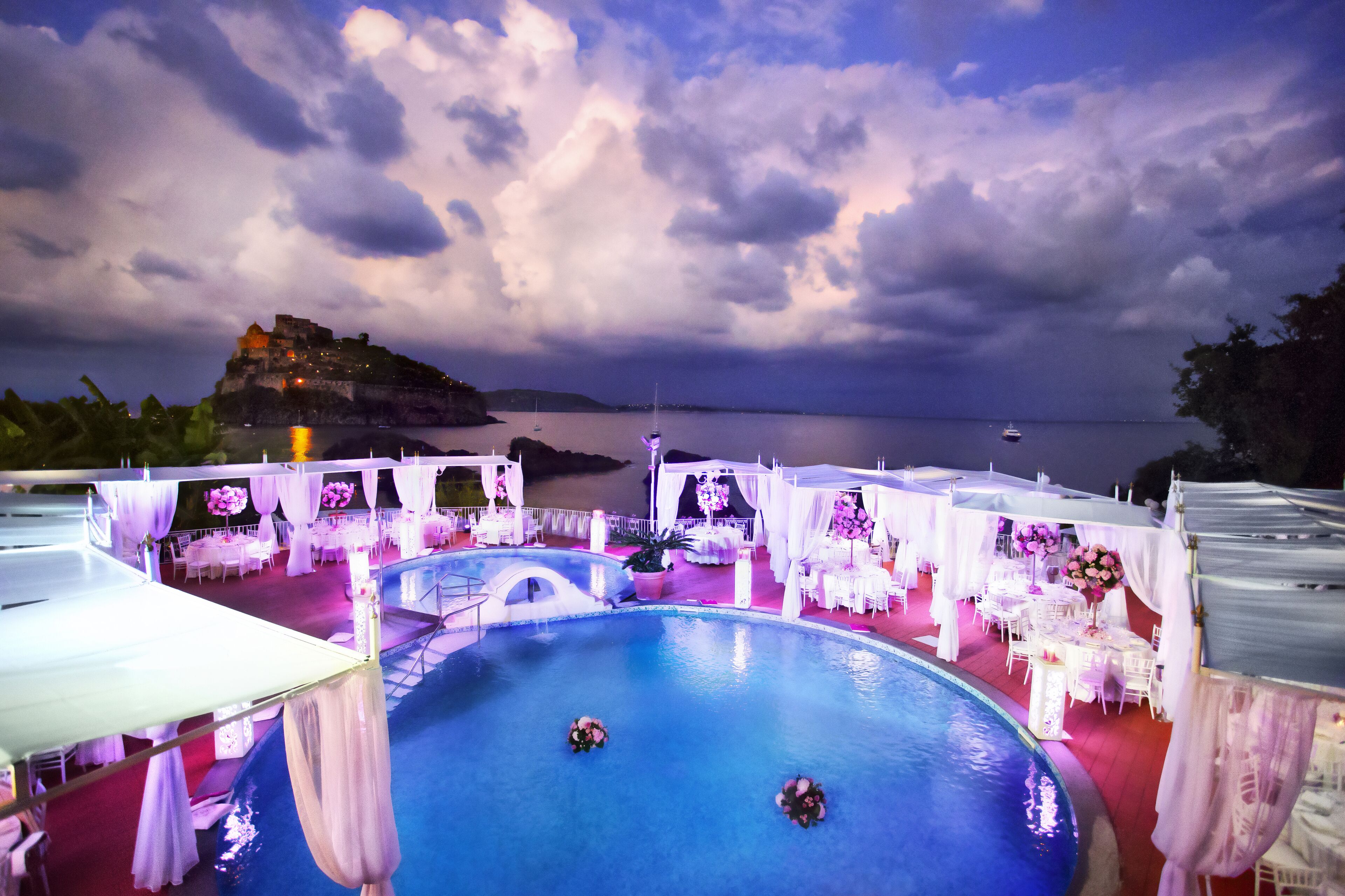 Un espace aménagé au bord de la piscine, avec des tables et des chaises, une piscine, et une vue sur la mer et une île lointaine.
