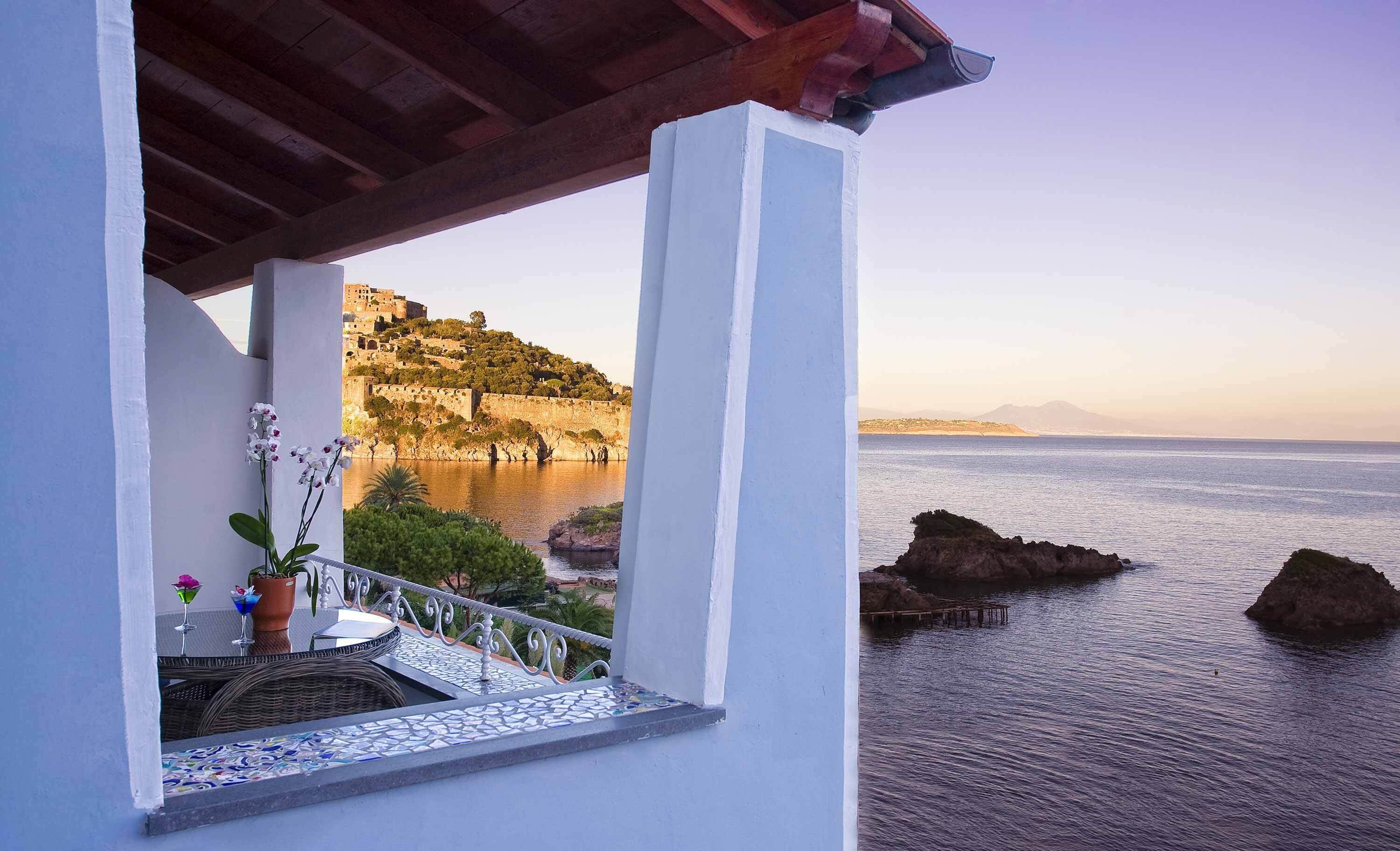 Un balcón con vista a una ciudad costera, que incluye un castillo histórico en una colina y el mar.