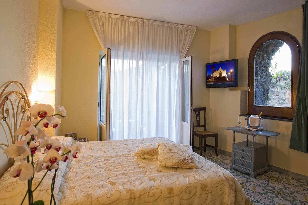 A hotel room with a bed, a small table with a bottle and glasses, a chair, and a TV on the wall.