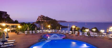 A pool area with lounge chairs and umbrellas, overlooking a historic building and the sea.