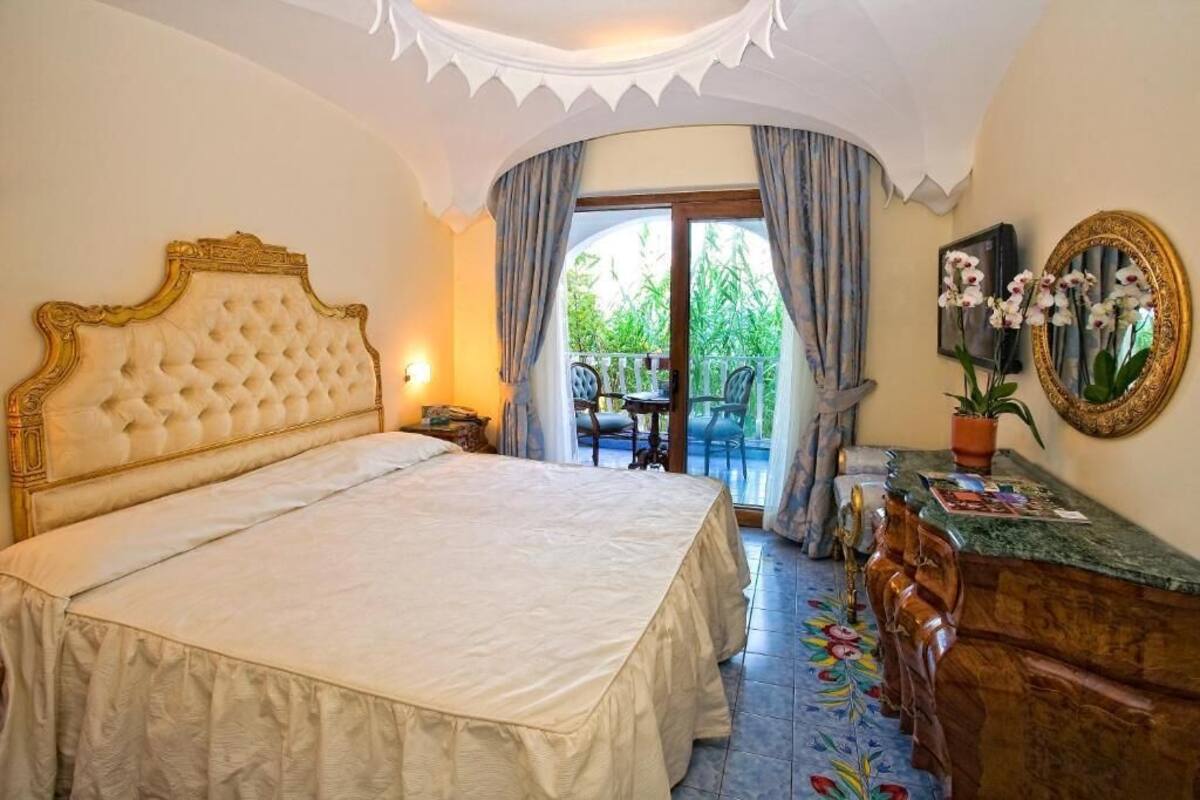A bedroom with a large bed, a wooden dresser, a mirror, and a balcony with a table and chairs.