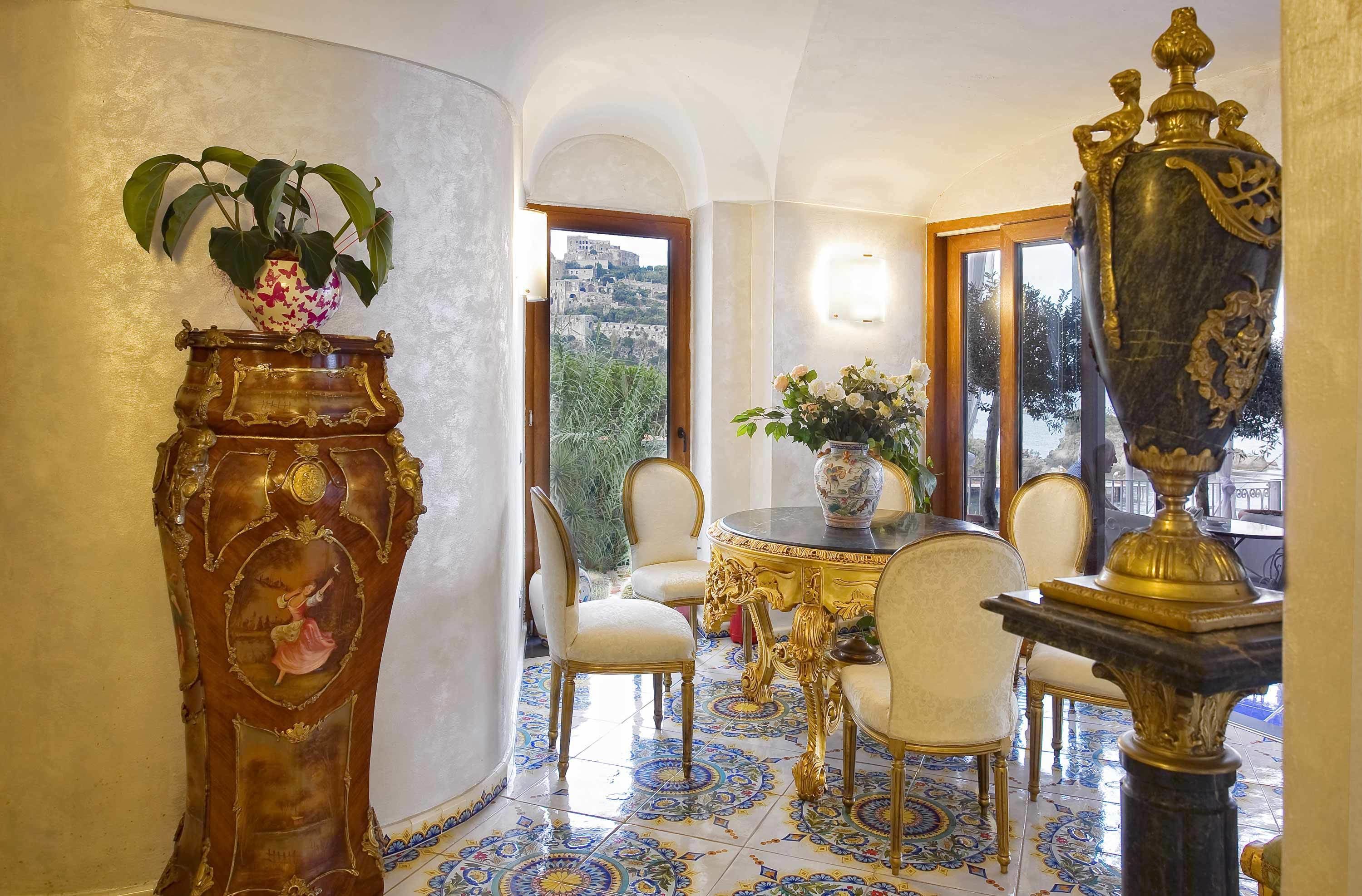Una habitación con una mesa redonda, sillas blancas, un jarrón grande y ornamentado, y una planta en maceta.