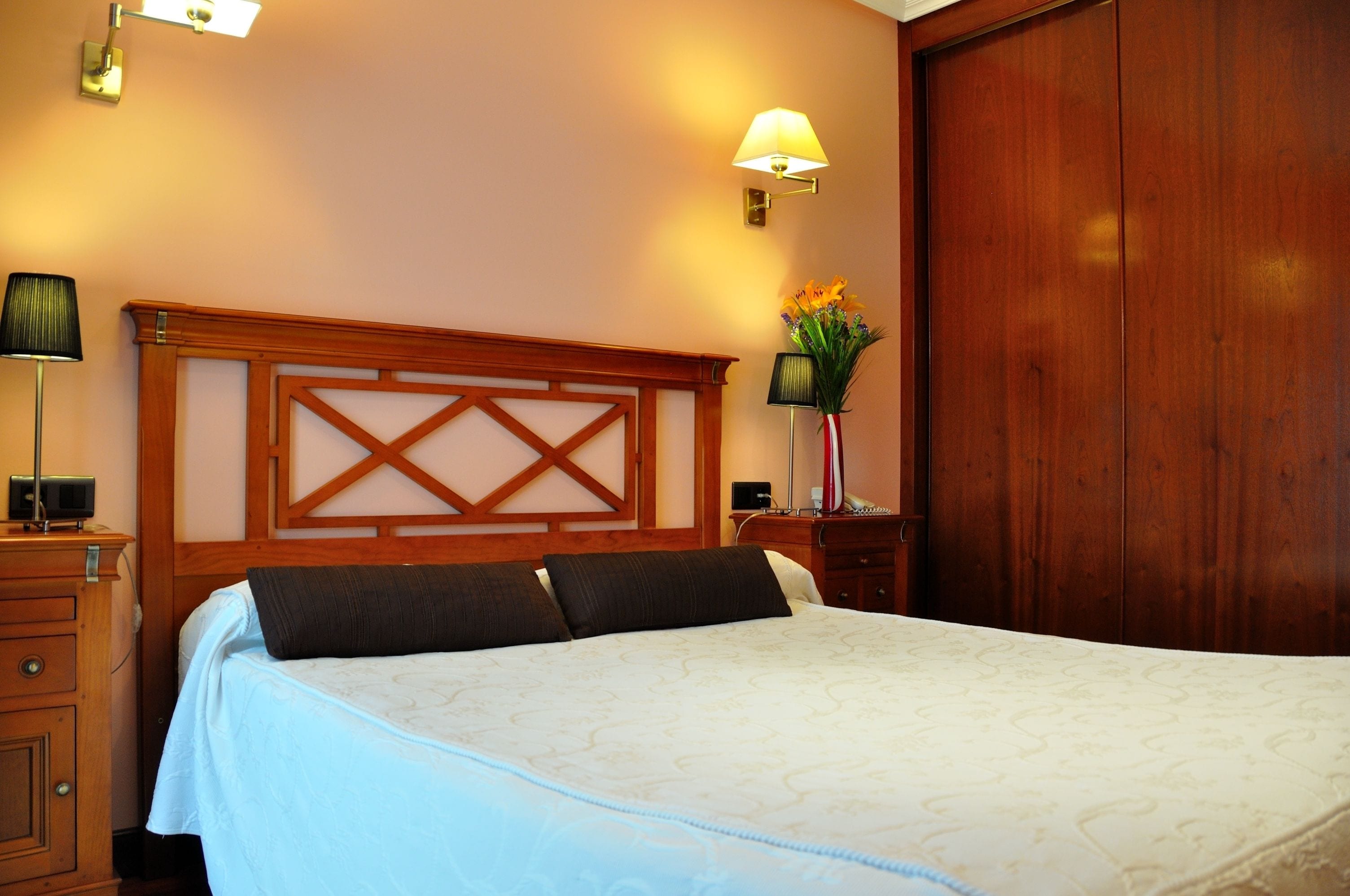 A hotel room with a large bed, wooden headboard, bedside lamps, a nightstand, and a wooden wardrobe.