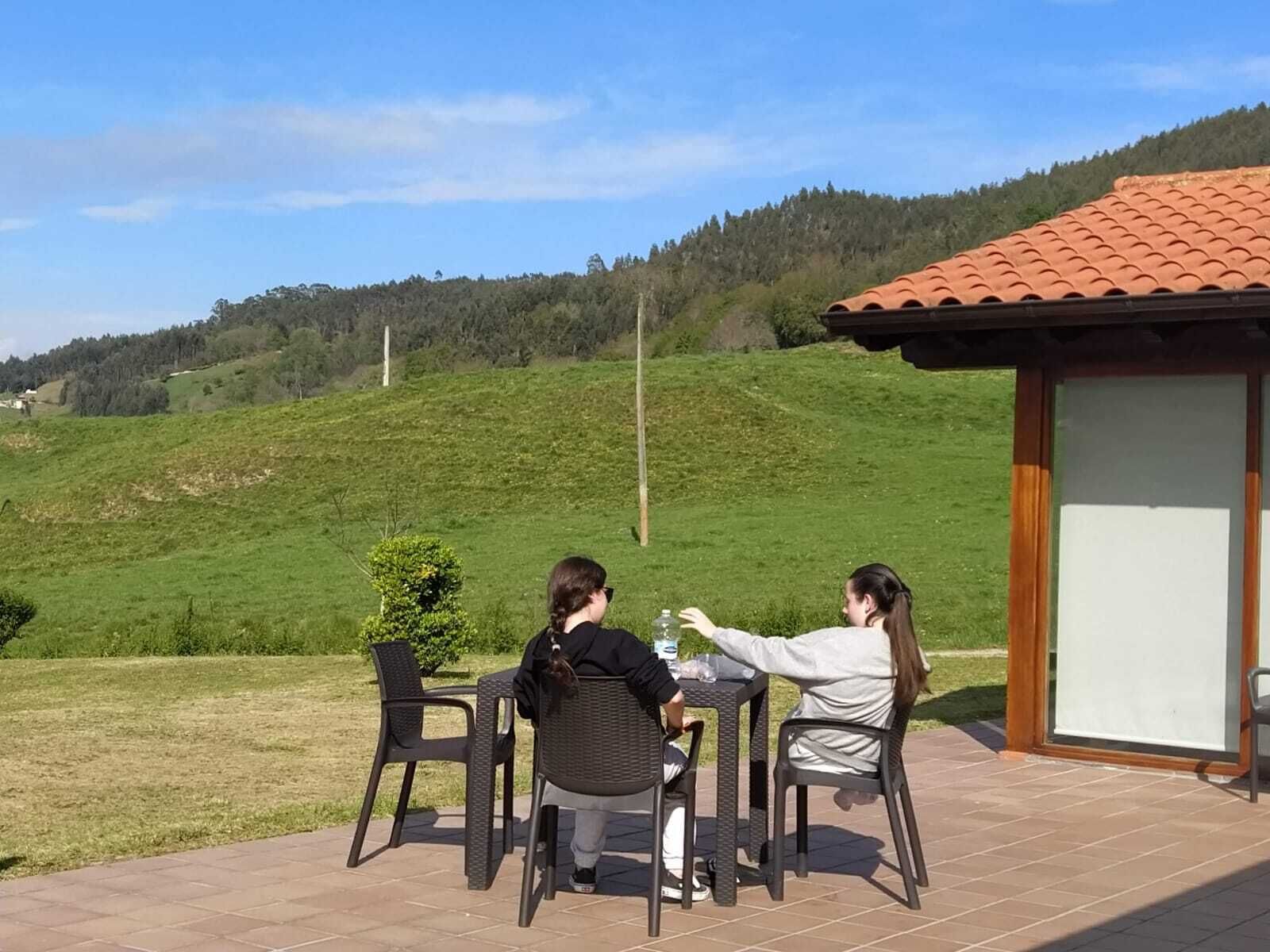 Twee personen zitten aan een buitentafel met uitzicht op een groene heuvel en een gebouw met een pannendak op de achtergrond.