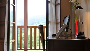 A room with a view of a green landscape through a large window, a desk with a laptop, a vase with a flower, and a standing lamp. - Hotel Puerta del Oriente (Llanes)