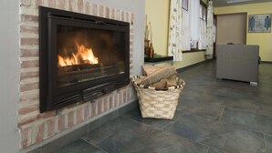 A fireplace with a brick surround, a woven basket with logs, and a wicker chair in a room with tiled flooring and a yellow wall. - Hotel Puerta del Oriente (Llanes)