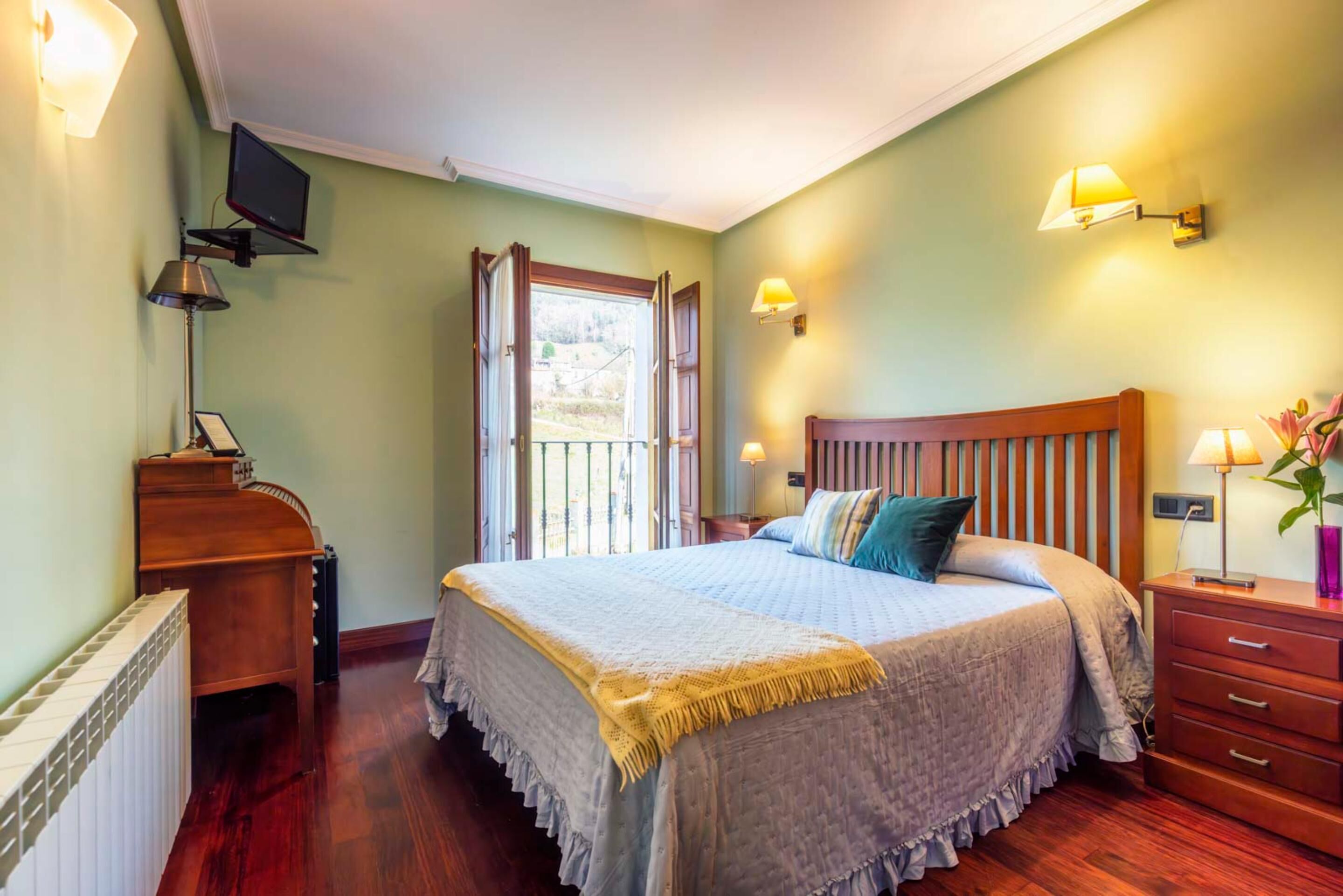 A bedroom with a wooden bed, a nightstand, a desk, a television, and a window with a view of the outdoors.