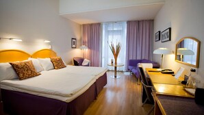 Habitación de hotel con cama, escritorio, silla, mesita y ventana con cortinas.