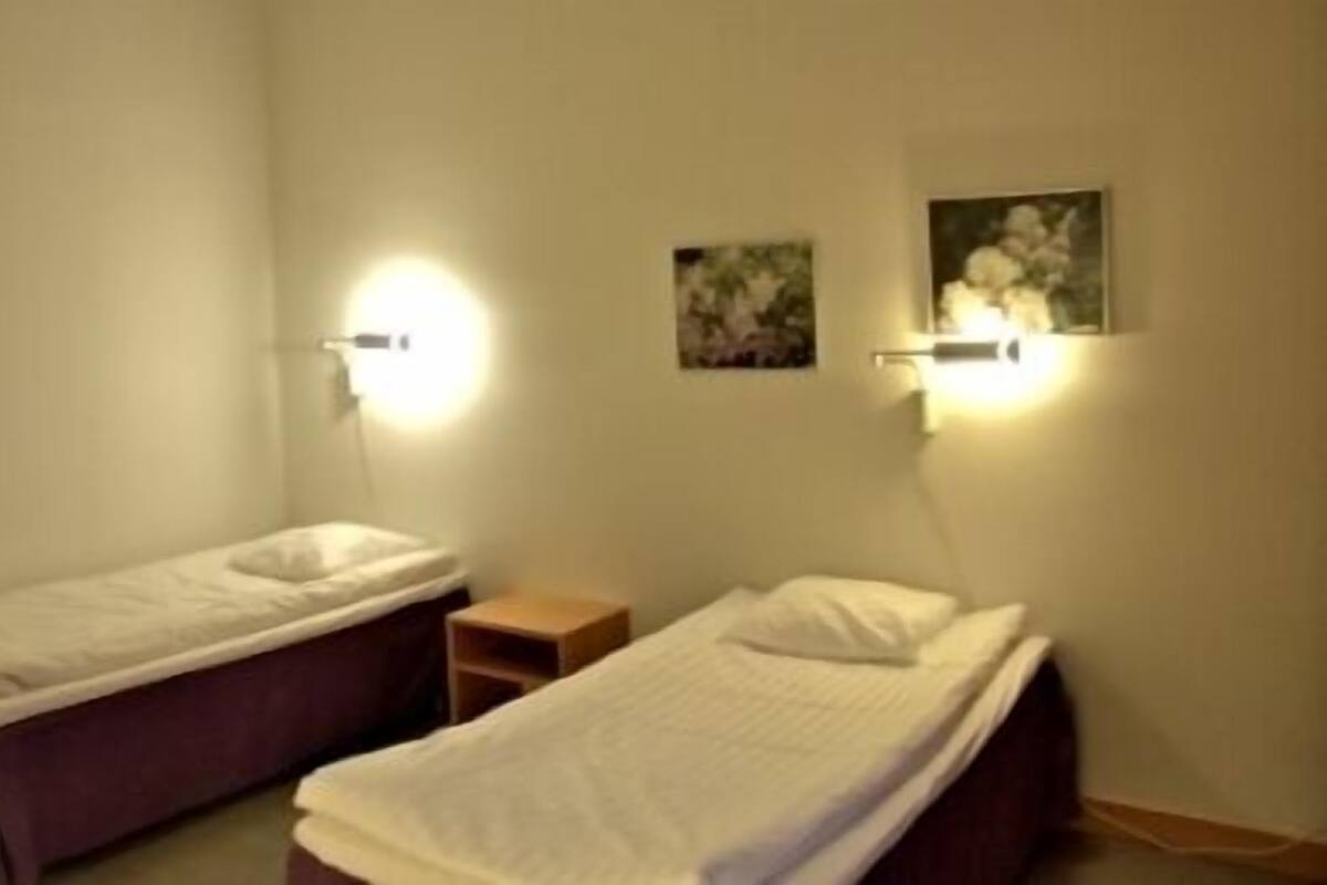 A small, dimly lit room with two single beds, a nightstand, and wall-mounted lights.