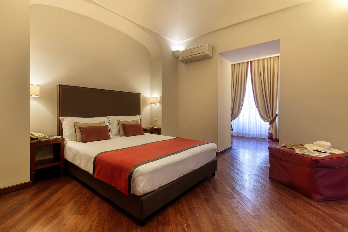 Ein Hotelzimmer mit einem großen Bett, Holzboden, einem roten Pouf und Blick ins Freie durch ein Fenster mit Vorhängen.