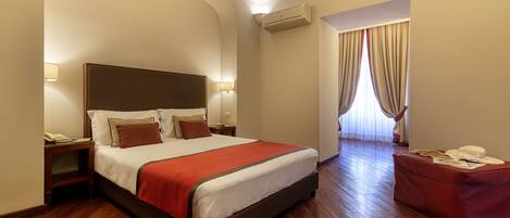 A hotel room with a bed, bedside tables, a red ottoman, and a view of the outdoors.