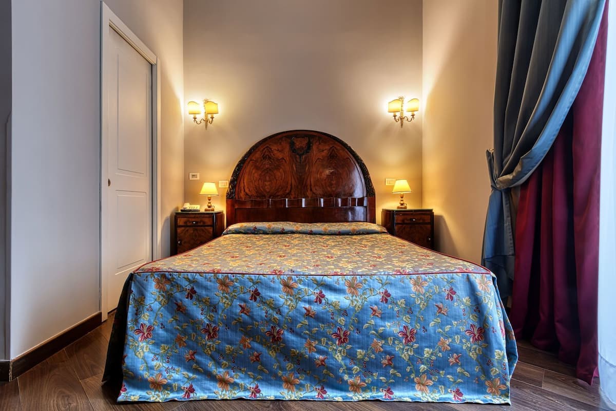 Ein Bett mit floralem Überwurf, zwei Nachttische mit Lampen sowie ein hölzernes Kopfteil.