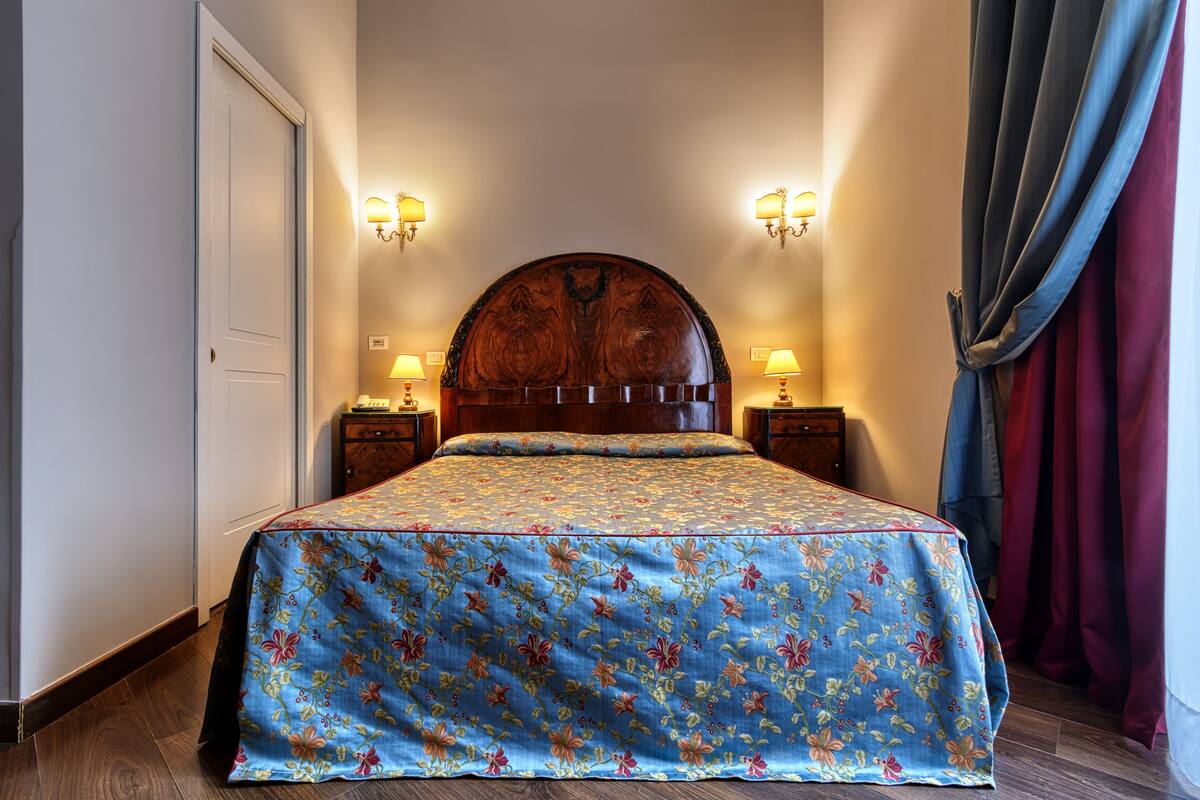 Ein Schlafzimmer mit einem Holzkopfende, zwei Nachttischen mit Lampen, einer blumigen Tagesdecke und Blick auf ein Fenster mit Vorhängen.