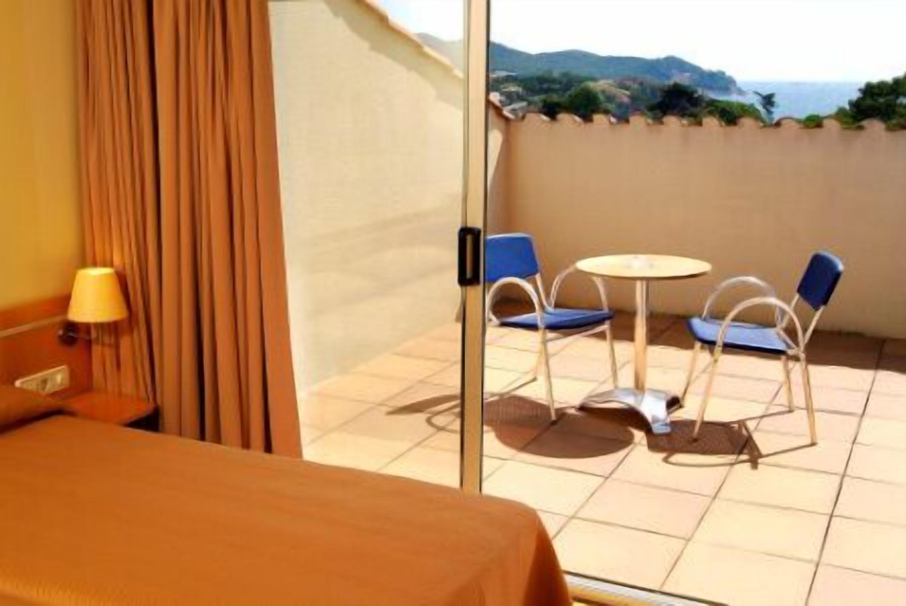 A hotel room with a bed, a table with two chairs, a view of the sea, and a curtain.