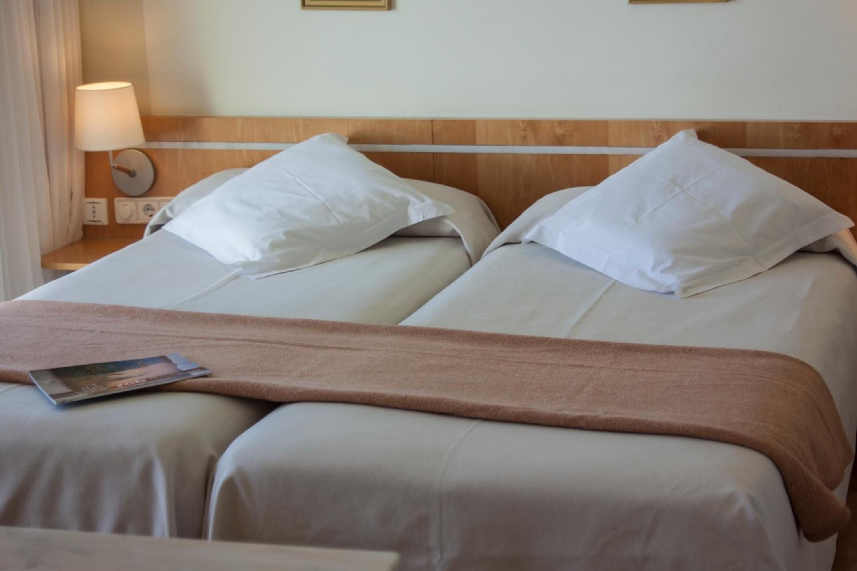 Et hotelværelse med to senge, hver med hvide hør og et brunt plaide. Der er et natbord med en lampe og et ur.