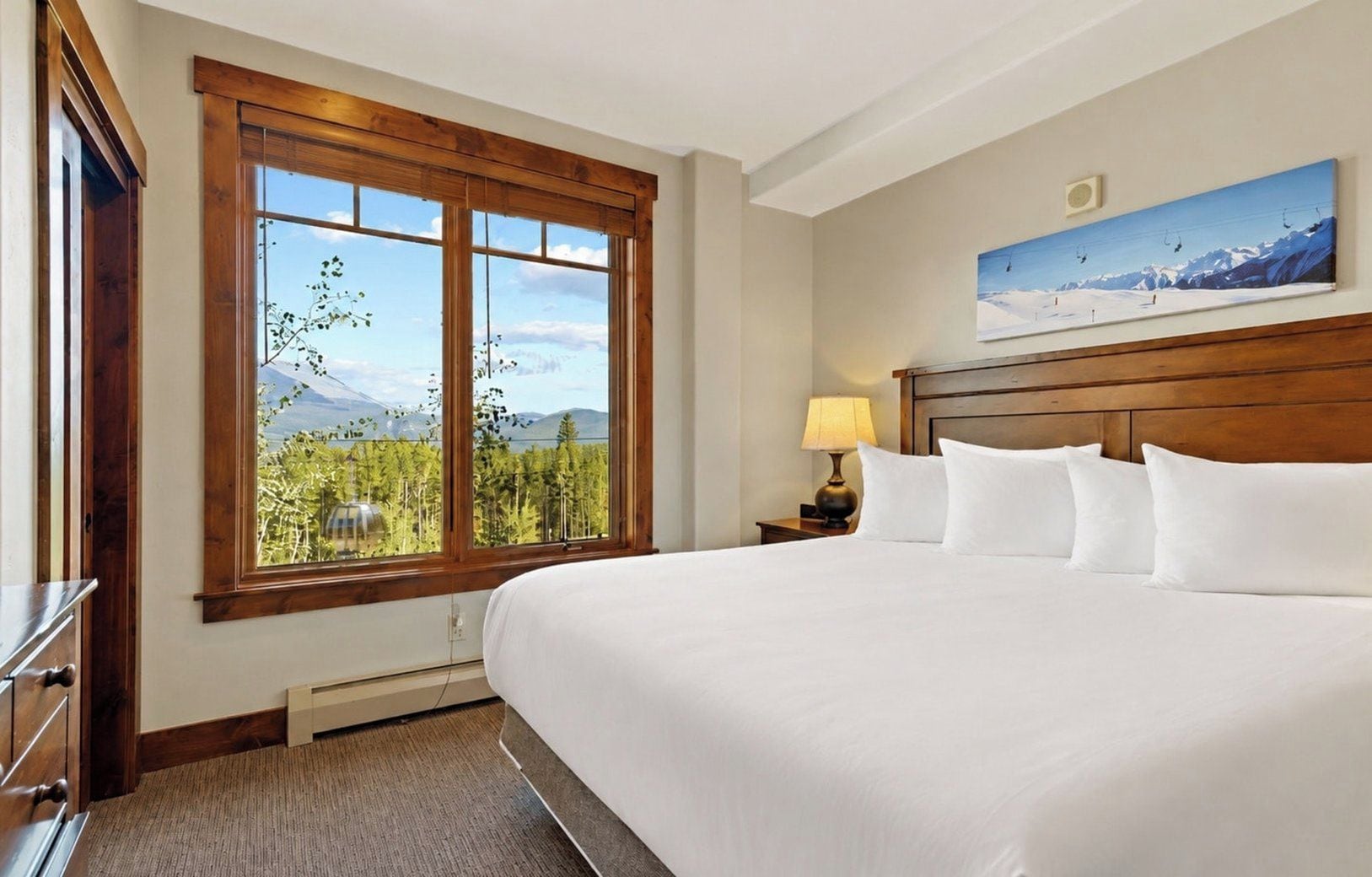 Een hotelkamer met een groot bed, een nachtkastje met lamp, een houten hoofdbord, een raam met uitzicht op bomen en bergen, en een schilderij aan de muur.