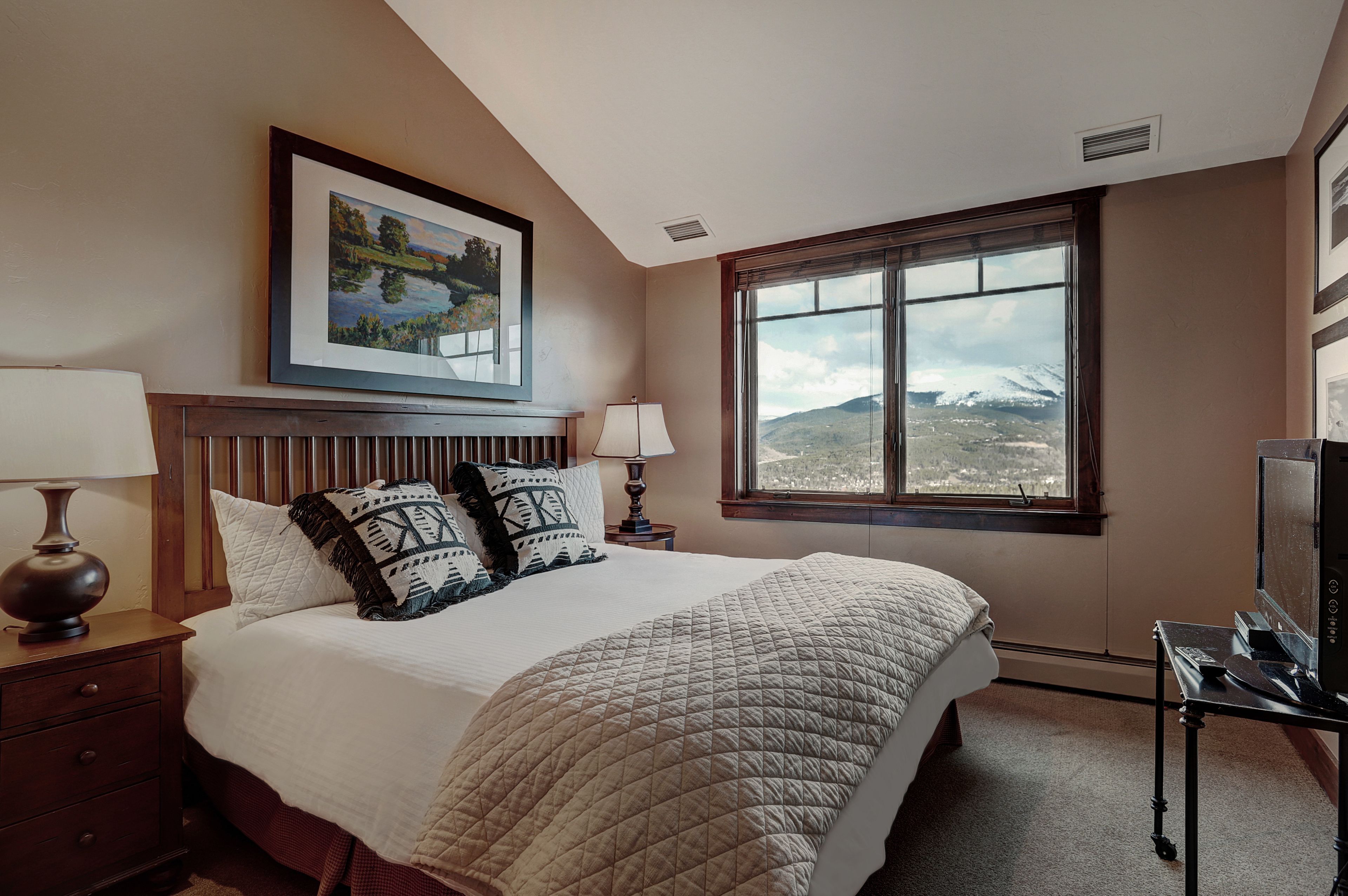 Une chambre à coucher avec un grand lit, un tableau représentant un paysage encadré, une fenêtre donnant sur les montagnes, une table de chevet avec une lampe et un téléviseur posé sur un meuble.