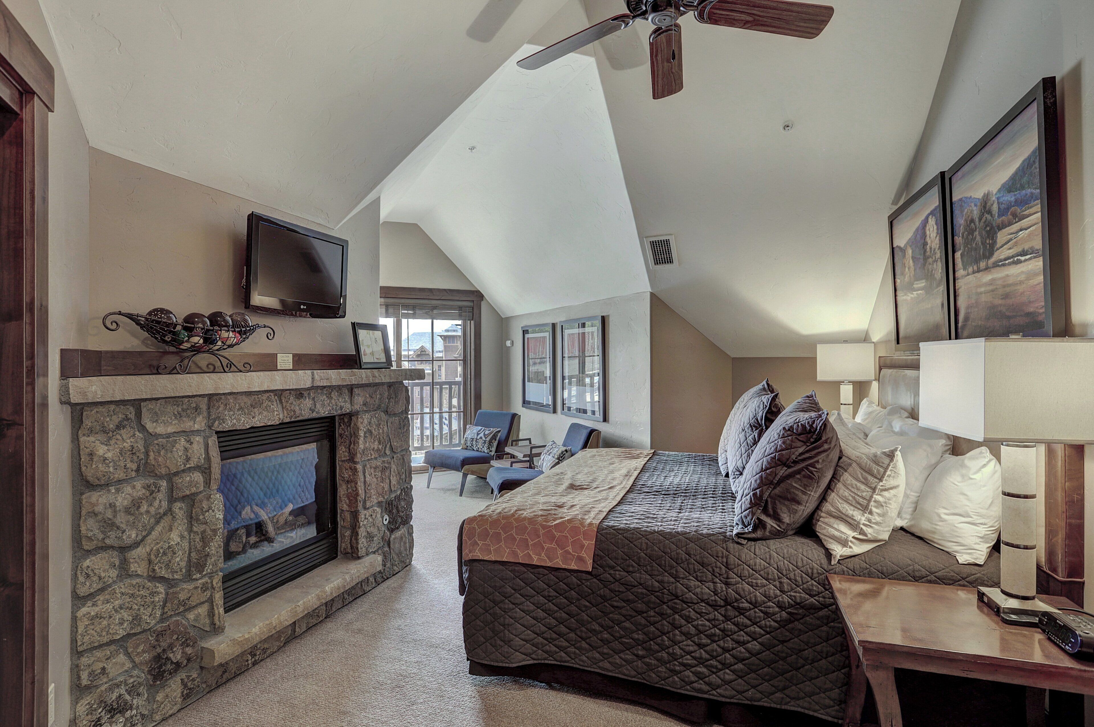 A bedroom with a stone fireplace, a bed with multiple pillows, a TV mounted on the wall, and a ceiling fan.