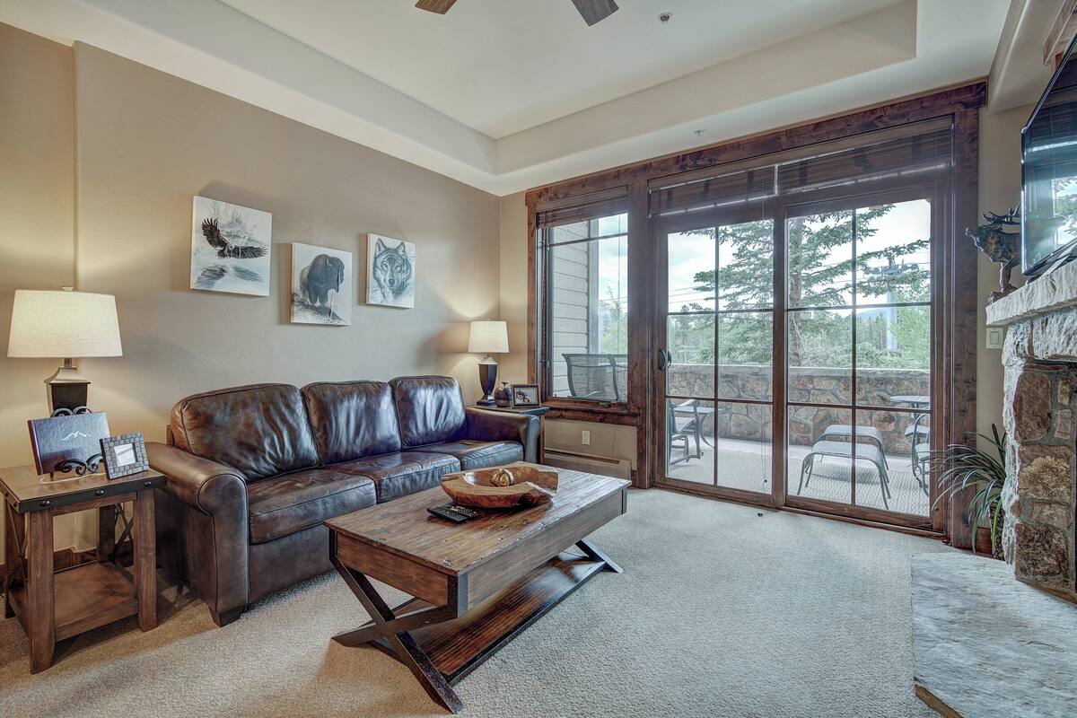 A spacious living room with a leather sofa, a wooden coffee table, and a fireplace. There are two lamps on side tables, and artwork on the walls.