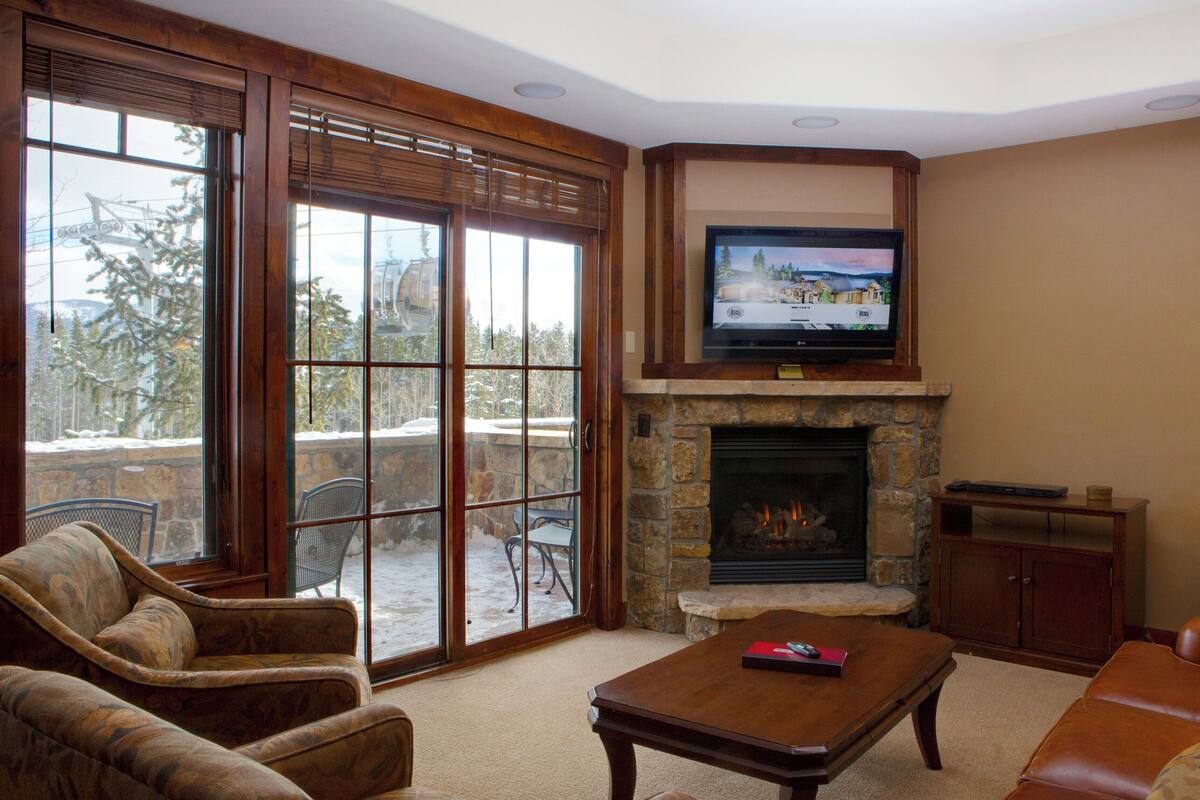 A cozy living room with a fireplace, a television mounted above it, and large sliding glass doors leading to a patio with a view of snow-covered trees.