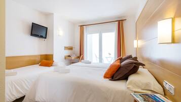 A hotel room with two beds, a television mounted on the wall, a window with curtains, and a small table with a lamp.