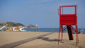 Una playa con una torre roja de salvavidas, un camino de arena y barcos al fondo.